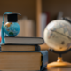 Ein kleiner Globus mit einer akademischen Doktorhut-Miniatur steht auf einem Stapel Bücher. Im Hintergrund sind ein klassischer Globus und Schreibutensilien in einer Bibliothek angedeutet.