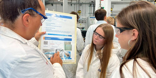 Wissenschaftlerin zeigt zwei Schulerinnen im Labor in Laborkitteln eine Tabelle im Labor.